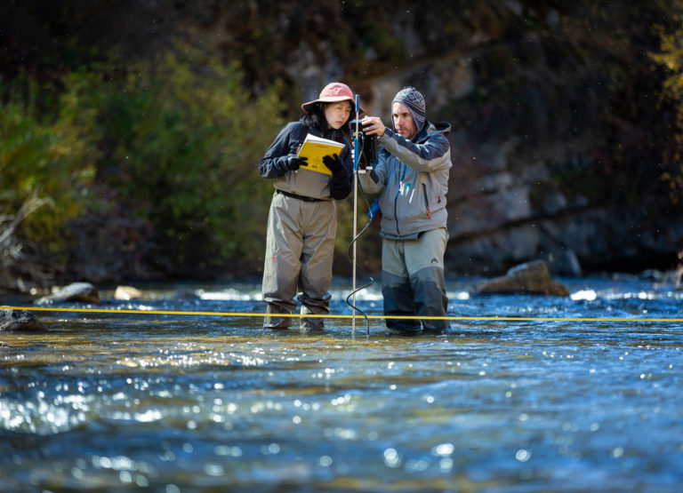 Two people in waders and hats stand in the middle of a creek and measure depth