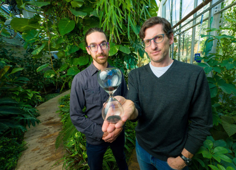 Peter Adler and PhD student Michael Stemkovskient stand in a greenhouse filled with plants and hold a hourglass