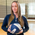 Former volleyball player Amy Crosbie works on equity issues in women's sports these days. She poses in the gym with a volleyball and net.