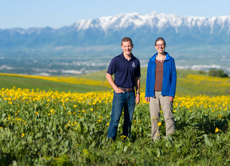 Jennifer Reeve, associate professor of organic and sustainable agriculture, and Matt Yost, associate professor and agroclimate Extension specialist, stand in a field of yellow flowers with the snowcapped Wellsville Mountains in the background.