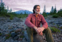 Charlie Crisafulli sits on a rock amidst pine trees at sunset with Mount St. Helens rising behind him in the background.