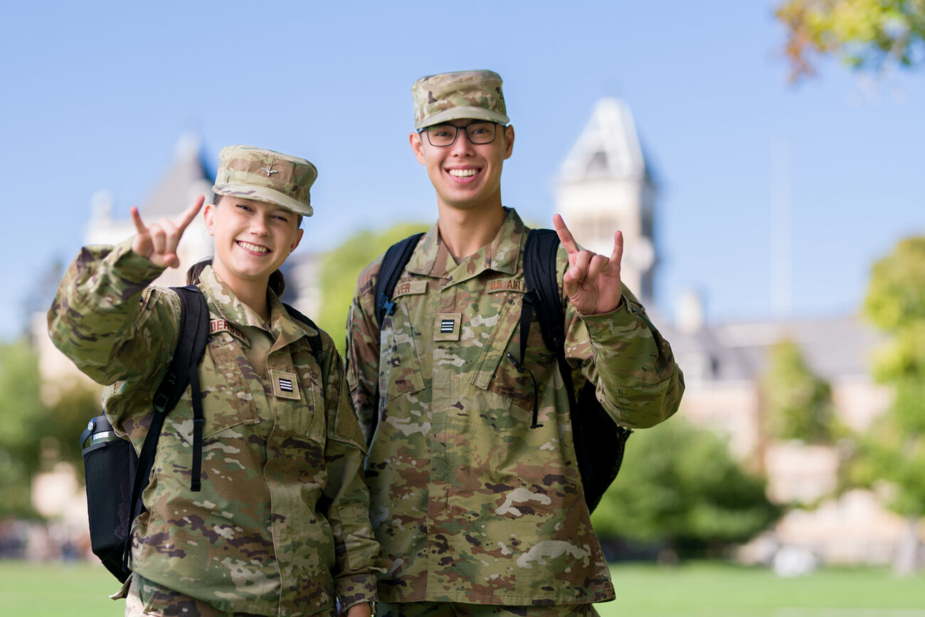 Tatum Nelson (right) throw's up USU's horns with his left hand while posing for a picture in fatigues with a fellow veteran on USU's Quad with Old Main in the background.