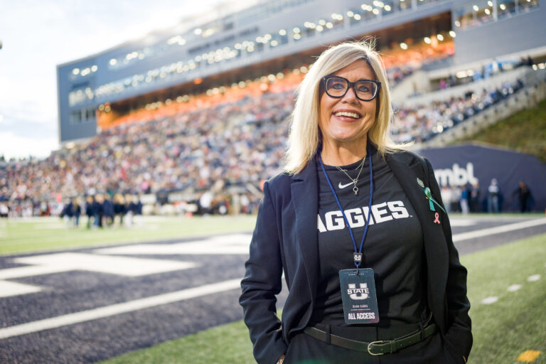 New USU athletic director Diana Sabou poses for a picture in the north end zone of Maverik Stadium, with USU's West Stadium Complex and press box rising behind her in the background.