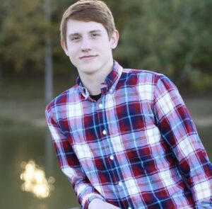 Cason Anderson poses for a portrait in a flannel shirt in front of a blurry lake and trees in the background.