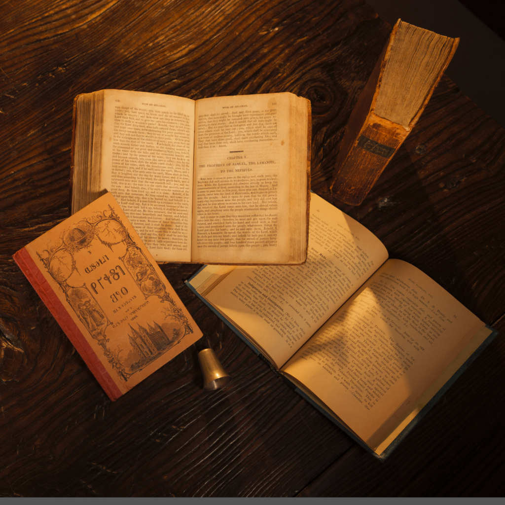 Two original copies of The Book of Mormon, a Deseret Reader, and other Latter-day Saint historical artifacts sit on a table.