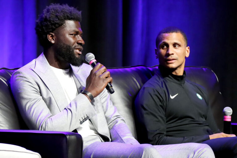 Two men sit on a couch with on holding a microphone speaking at an event.