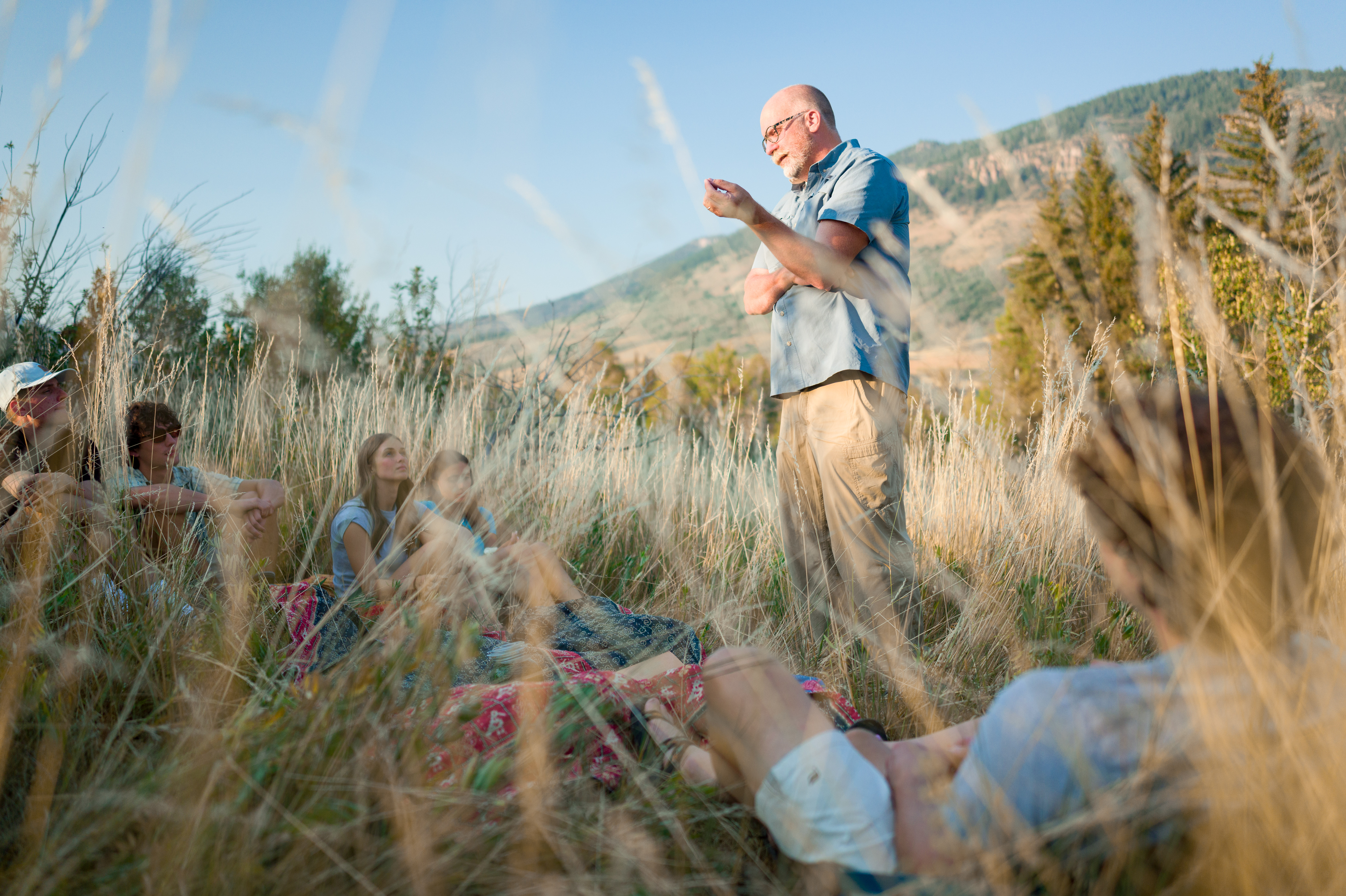 A older professor stands in knee-high grass speaking to a group of sitting students in an outdoor setting.