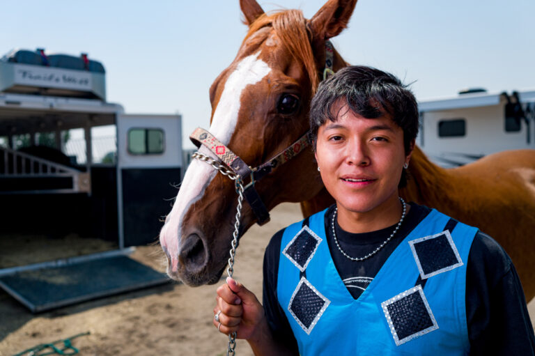 A Native American man stands holding a horse by the reigns.