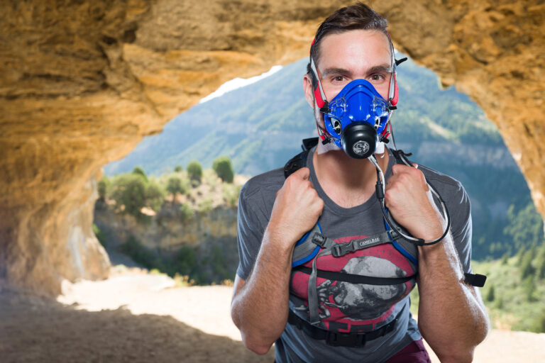 A man wearing what looks like a c-pap machine stands in front of the Logan Wind Cave after a research test.