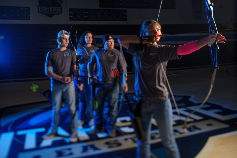 A woman pulls back a bow and poses for a picture in a gym with her teammates in the background.